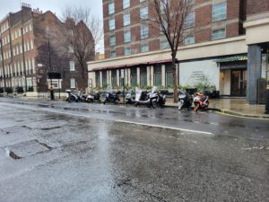Most motorbikes stand in the open rain while a cover protects one.